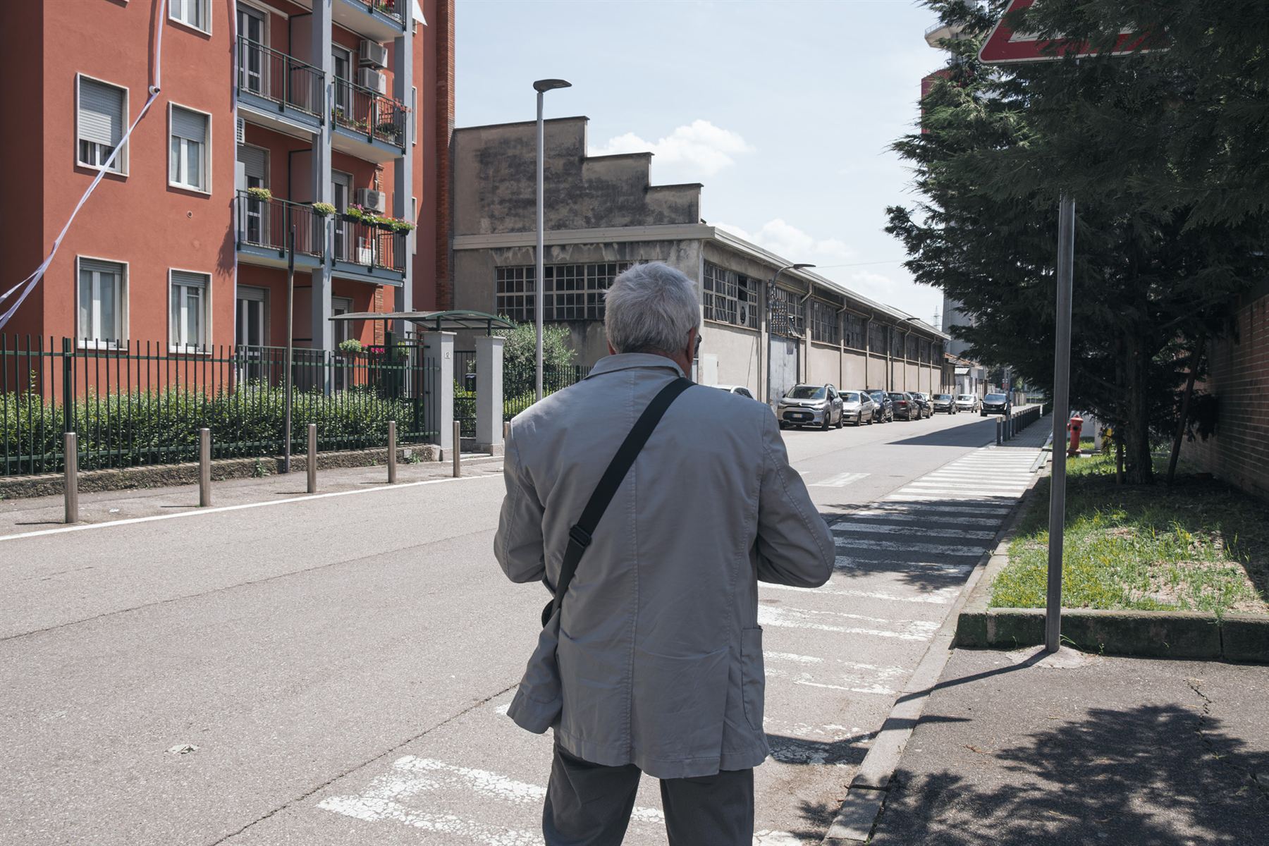 A pedestrian walking away from the camera along a residential street in Pero, between an apartment building and a former industrial building