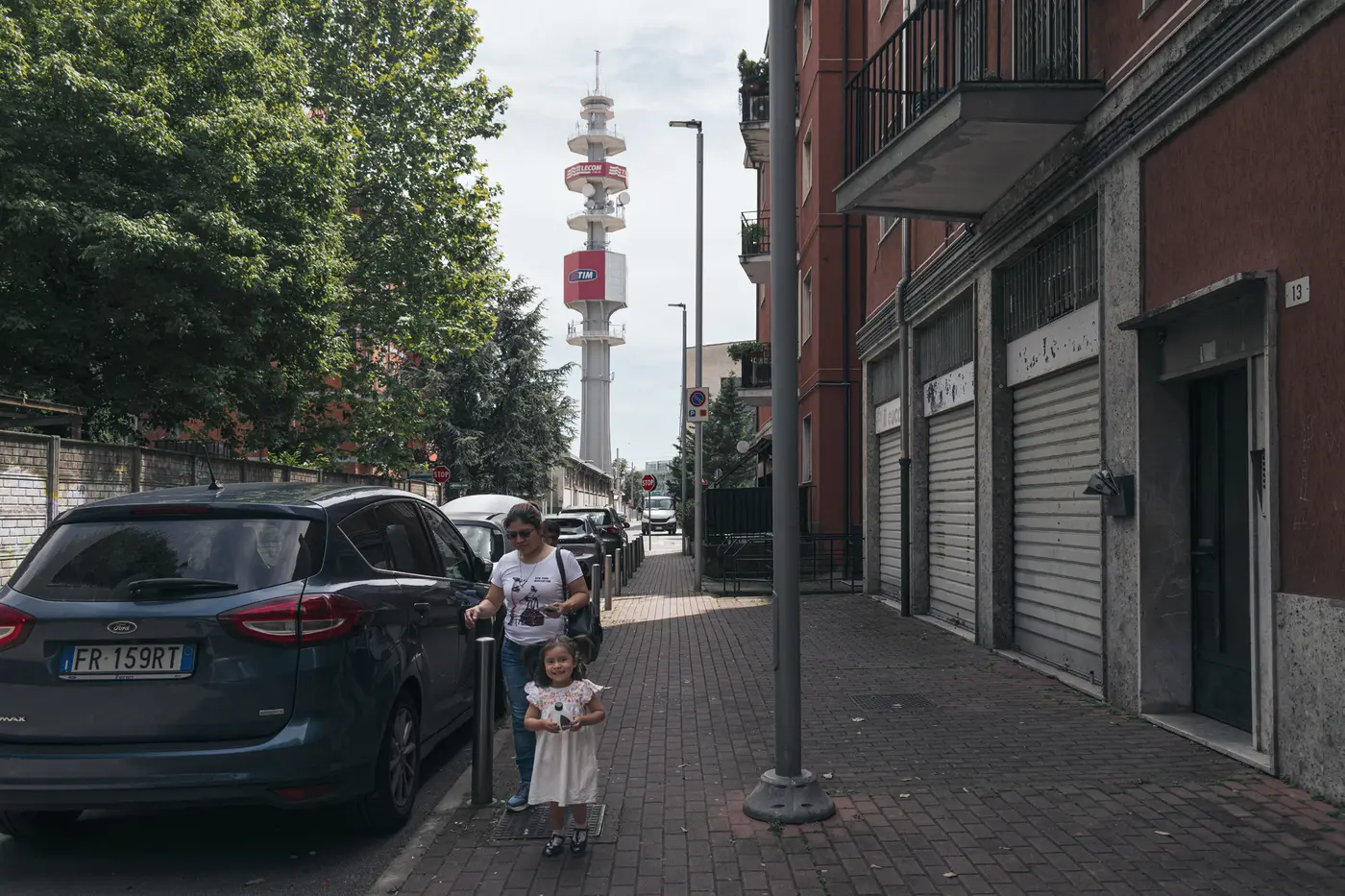 An adult and child moving along a narrow sidewalk between parked cars, shops, and a streetlight