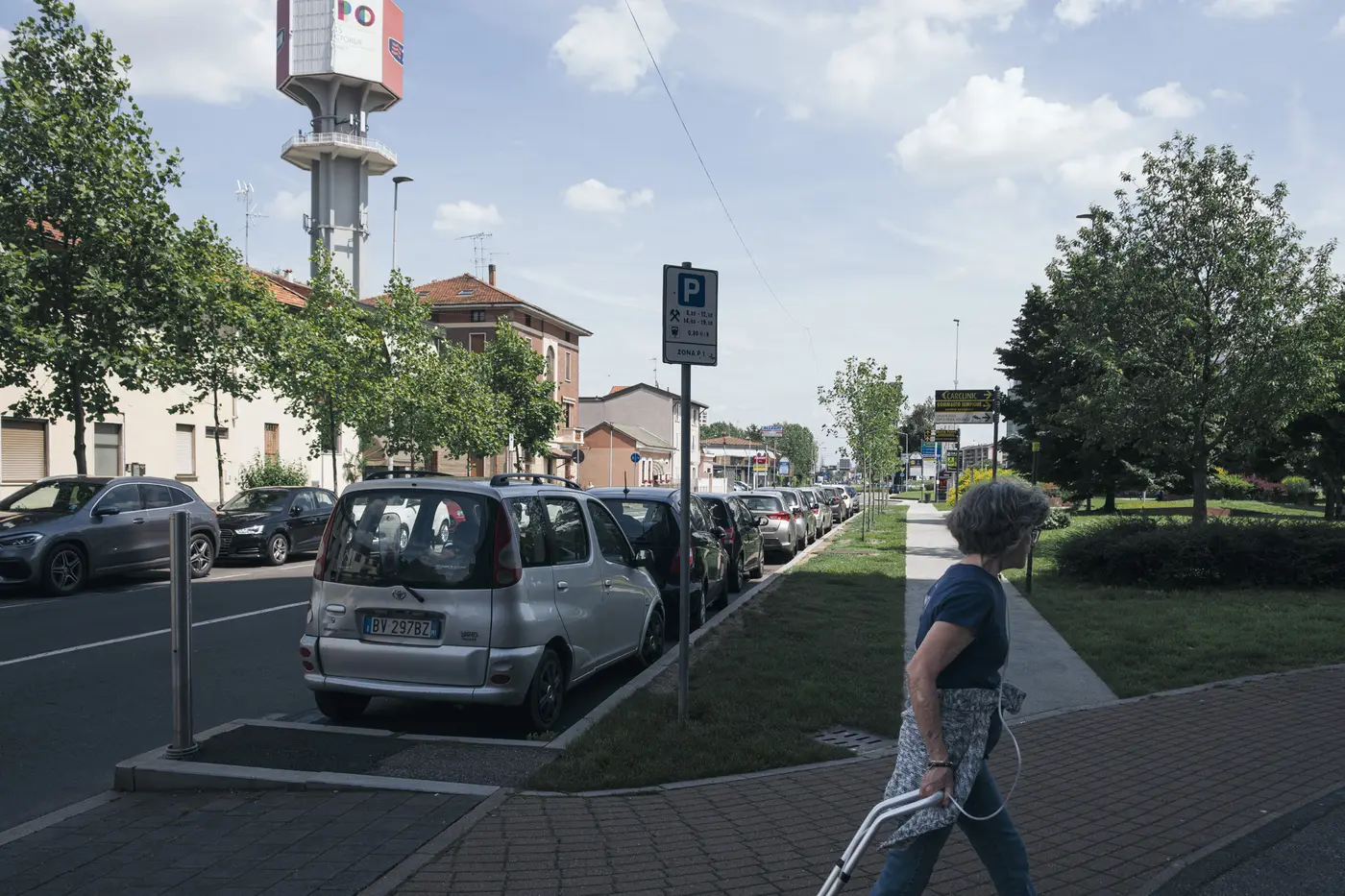 A pedestrian crossing near parked cars, housing, greenery, and Pero's water tower