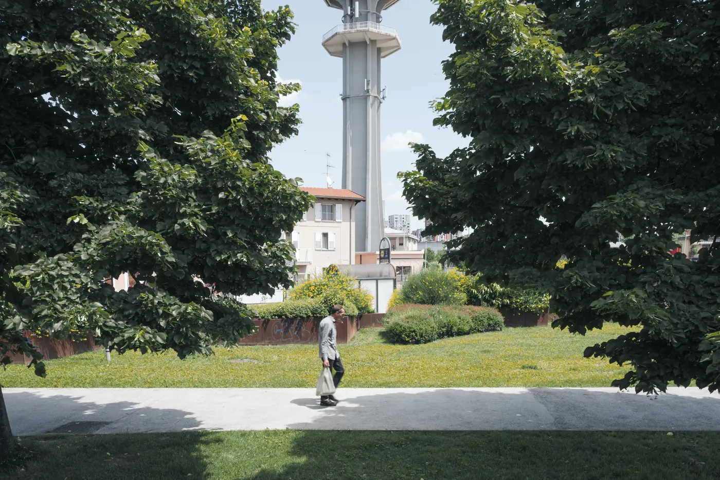 A lone pedestrian walking through a green space framed by mature trees and Pero's water tower