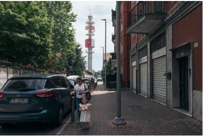 An adult and child moving along a narrow sidewalk between parked cars, shops, and a streetlight