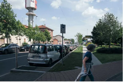 A pedestrian crossing near parked cars, housing, greenery, and Pero's water tower