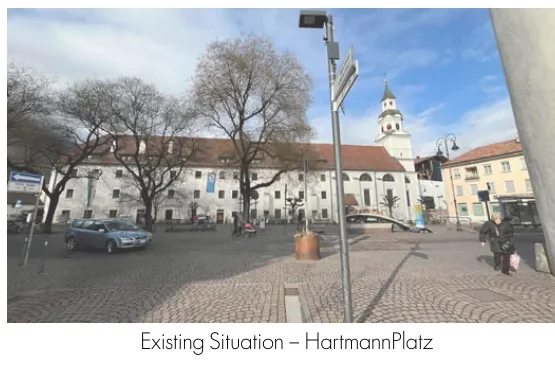Existing Hartmannplatz with parked cars, leafless trees, and the church beyond