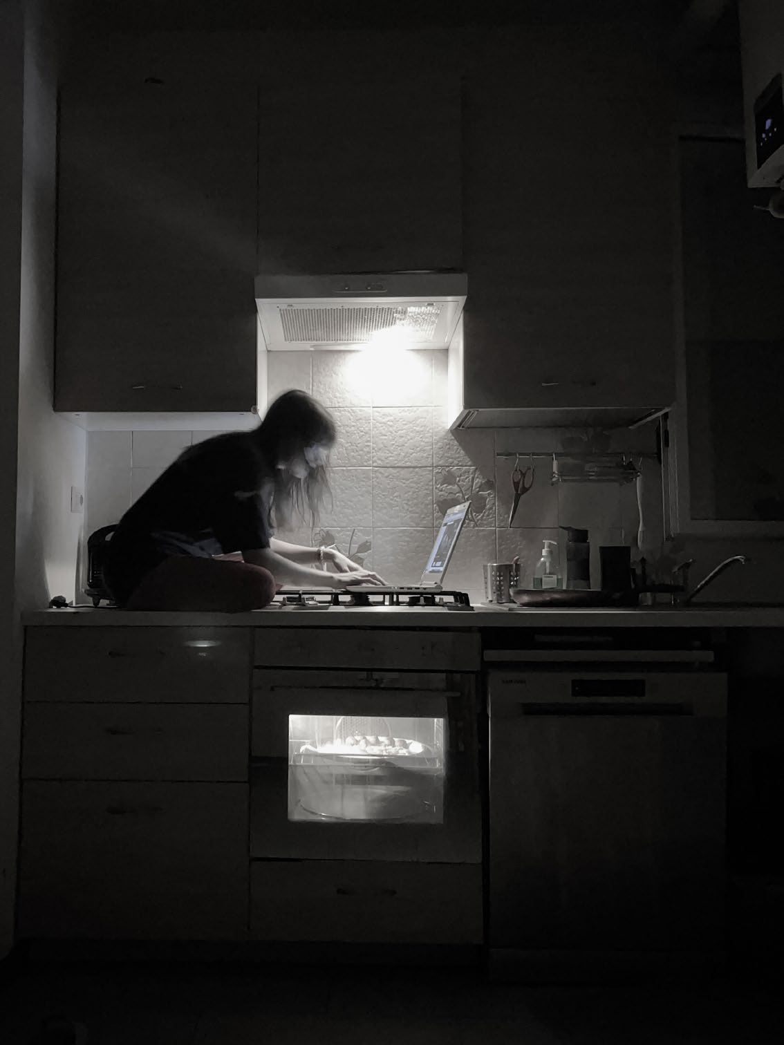 A solitary figure working in a dark kitchen, illuminated by the light above the stove