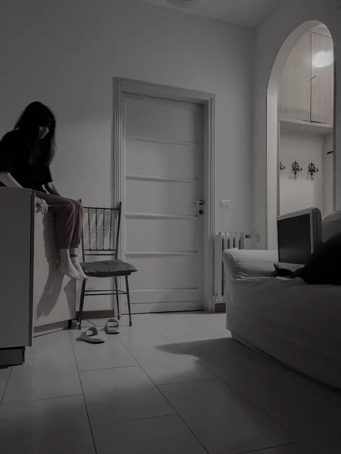 A dark-haired figure in a dark top sits barefoot on the edge of a kitchen unit beside a wooden chair and a pair of slippers, with a white panelled door and an arched hallway behind