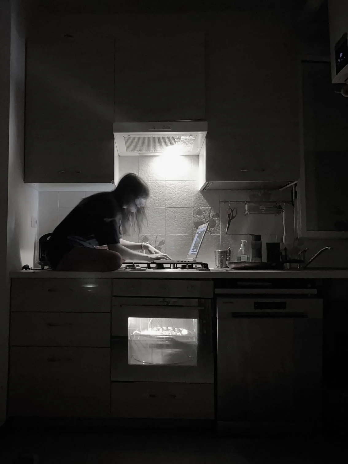 A solitary figure sits cross-legged on a kitchen counter working at a laptop, lit by the light of the cooker hood, with the oven glowing in an otherwise dark kitchen