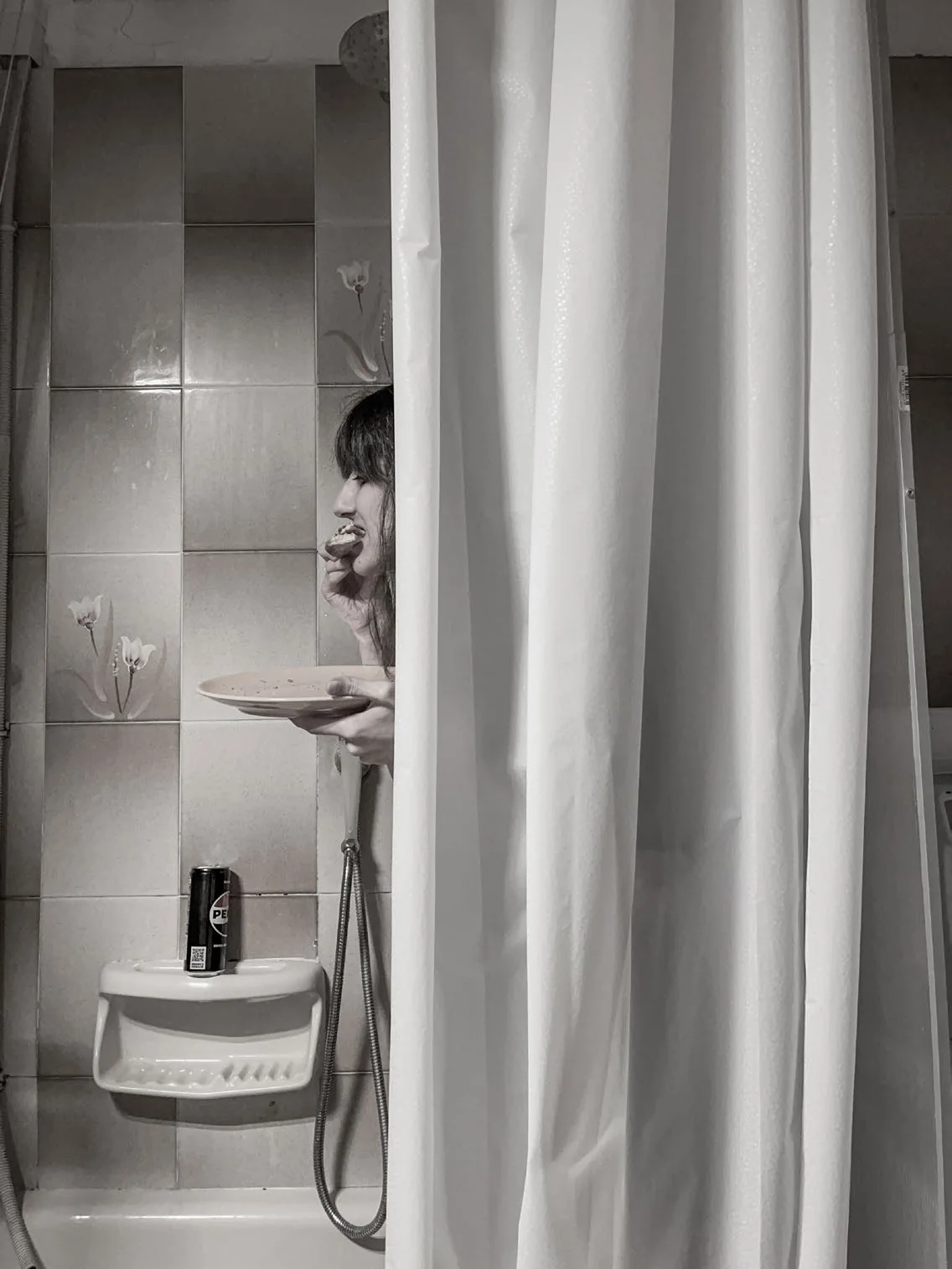 A figure eats from a plate inside a tiled shower, half hidden behind a white shower curtain