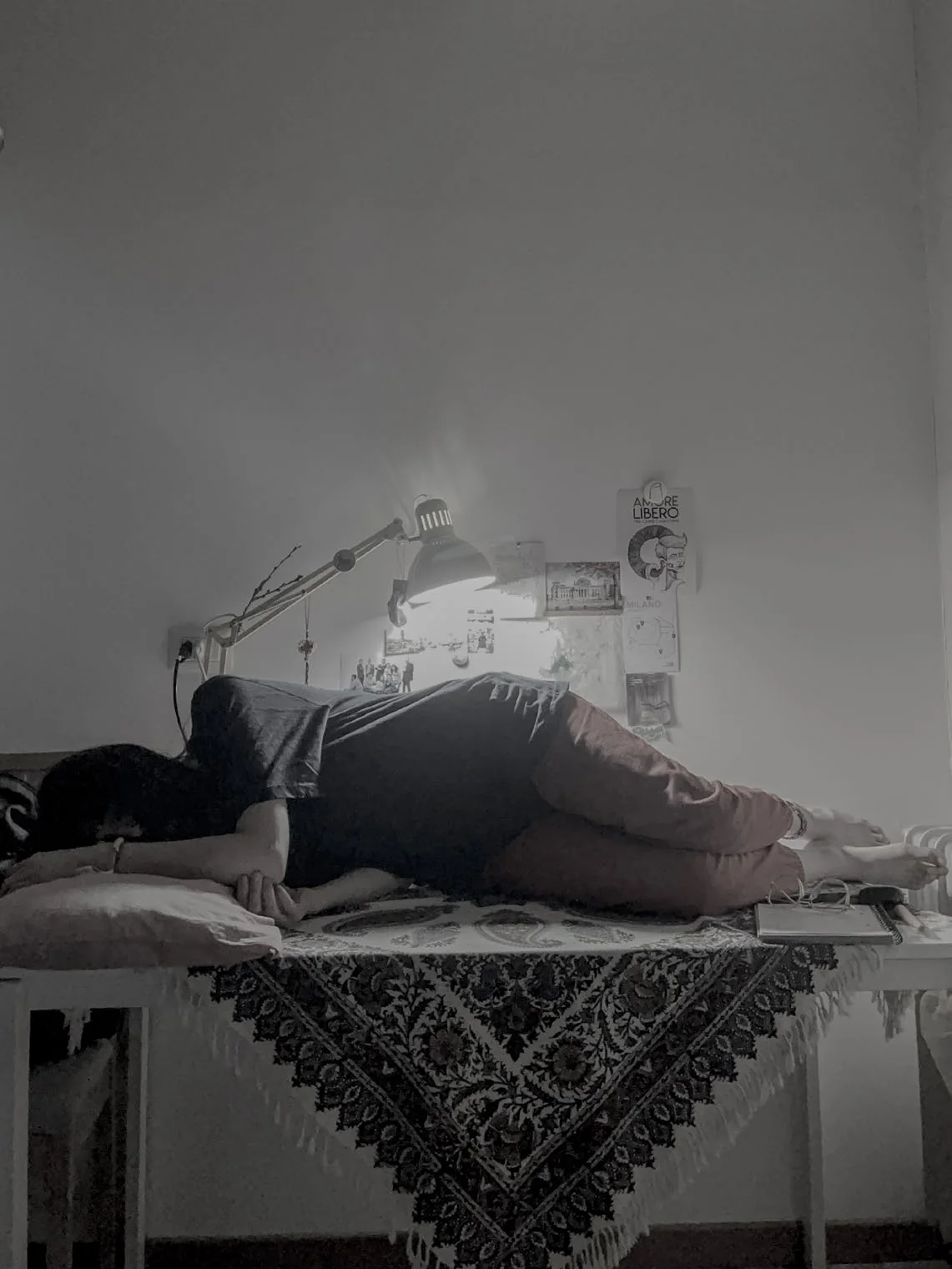 A figure lies curled on a table draped with a patterned cloth beneath an angled desk lamp, with postcards pinned to the wall behind