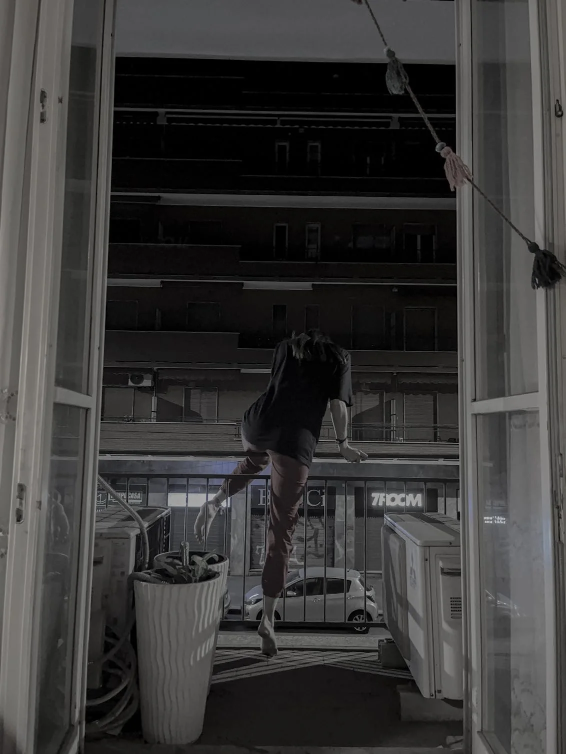 A figure, seen from behind, climbs over the railing of a balcony beyond open window doors, with a street of apartment buildings and a parked car beyond