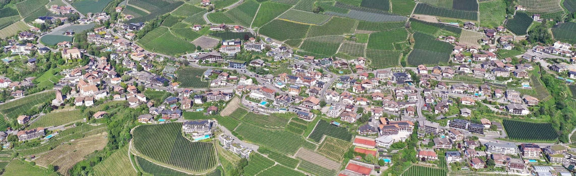 Panoramic aerial view of Schenna with a dashed square and a dashed circle marking two areas across the hillside